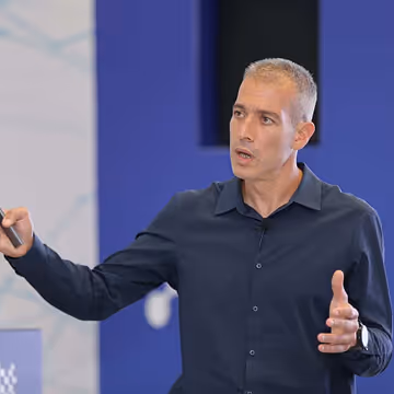 A man in a dark blue shirt giving a presentation with his left hand raised and right hand gesturing.