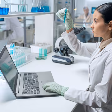 Female scientist in gloves examining a test tube with blue liquid while using a laptop in a laboratory.