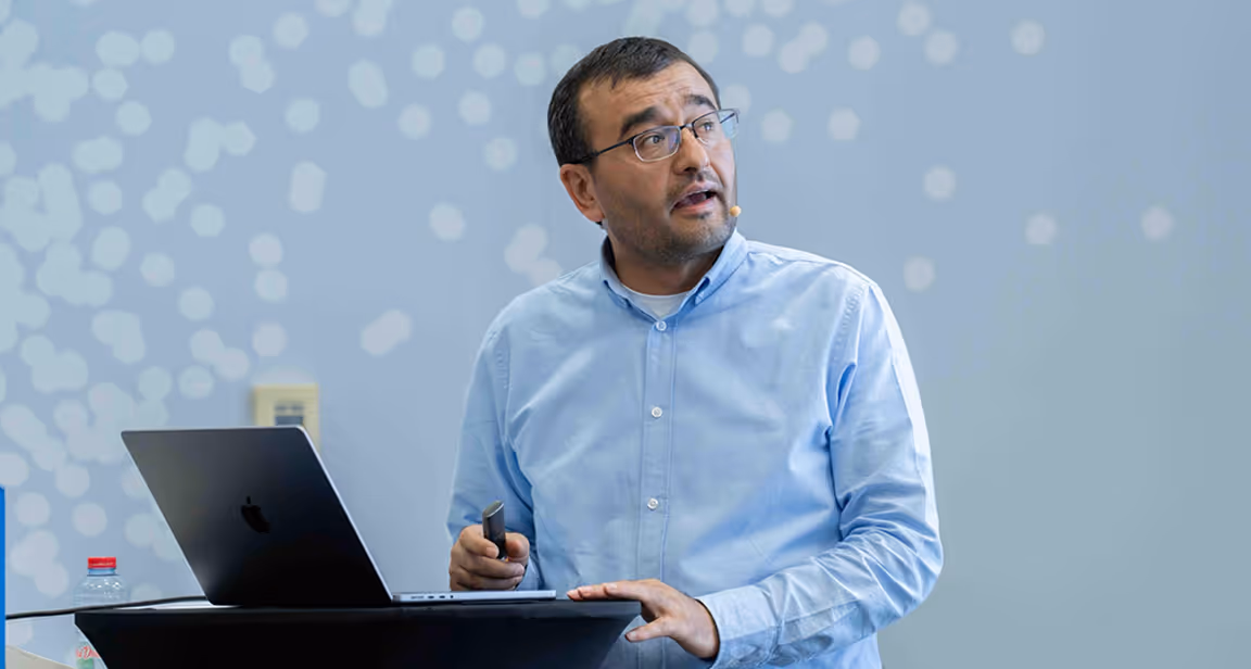 Man wearing glasses and a light blue shirt speaking at a podium with a laptop in front of him.