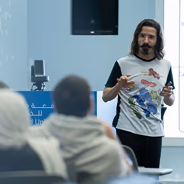 Man with glasses and long hair giving a presentation to seated audience in a modern classroom.