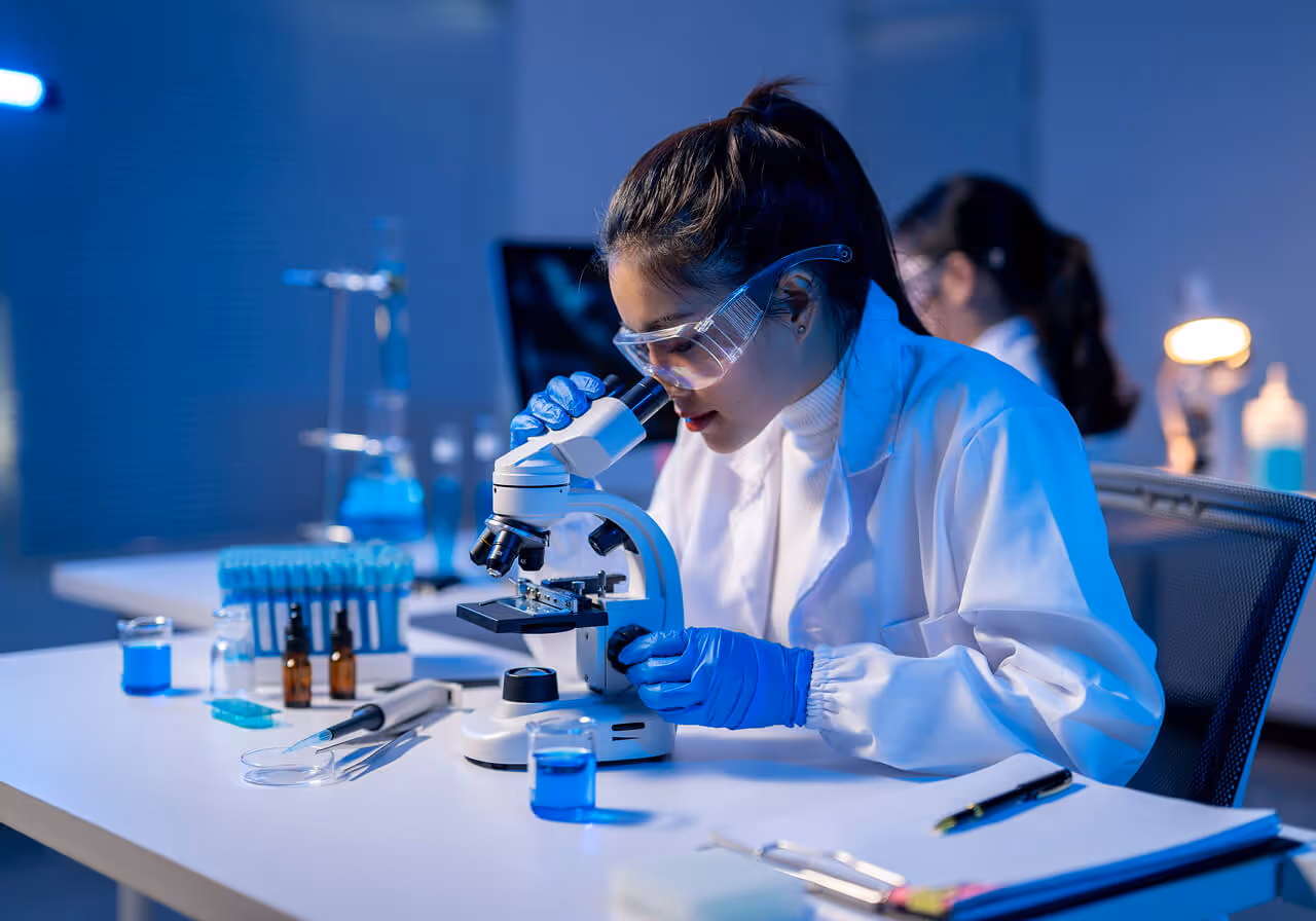 Female scientist wearing safety glasses and blue gloves examining a sample under a microscope in a laboratory with lab equipment on the table.